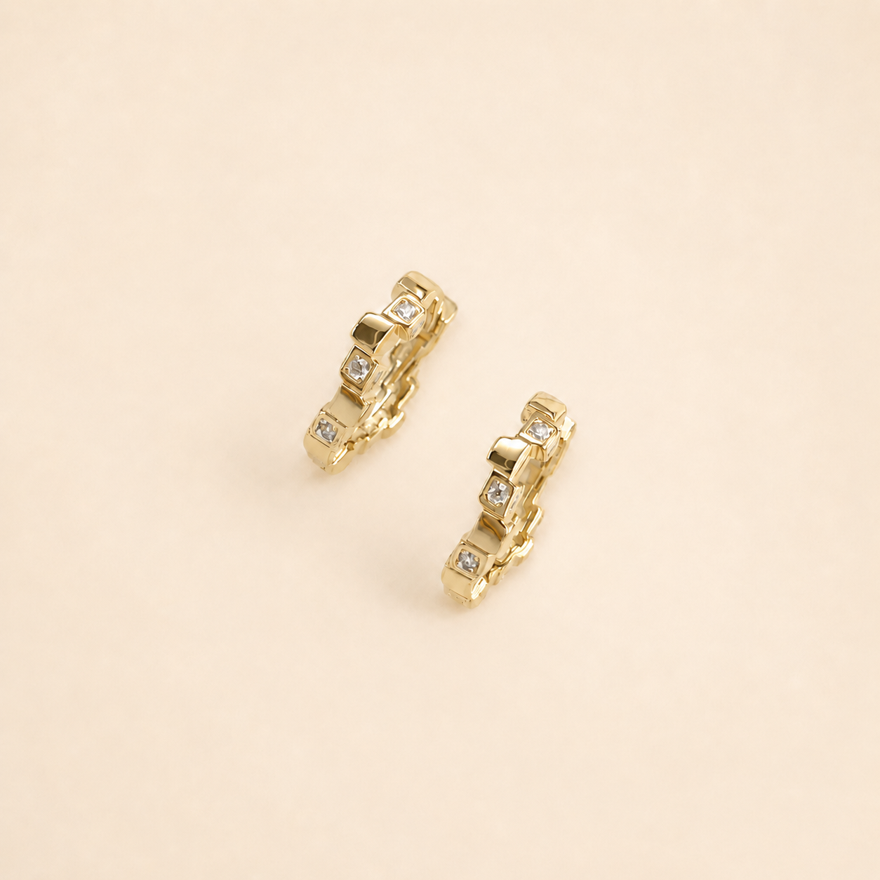 A pair of gold huggie hoop earrings with small clear gemstones, displayed on a solid beige background.