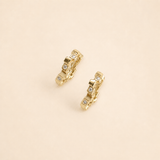 A pair of gold huggie hoop earrings with small clear gemstones, displayed on a solid beige background.