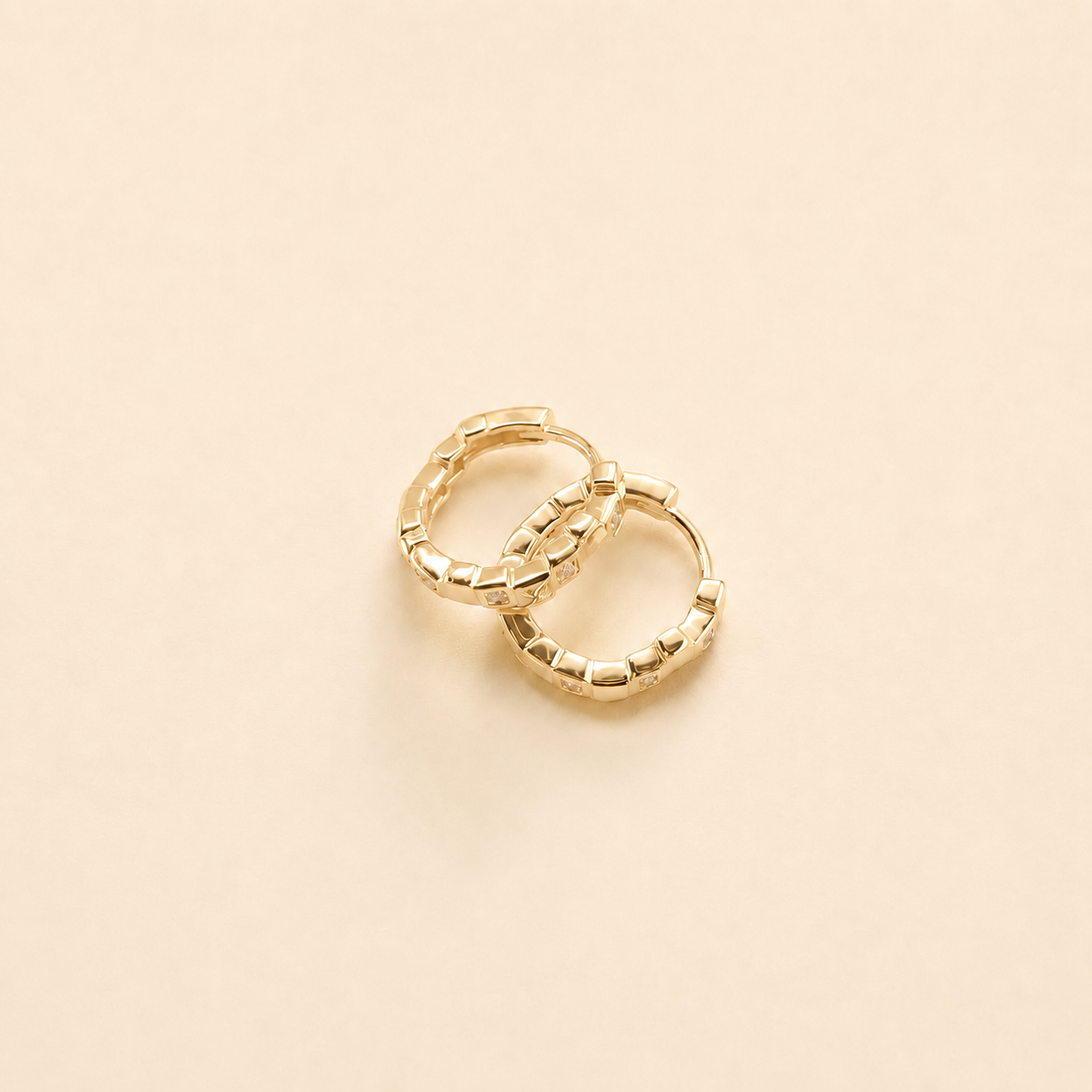A pair of small gold huggie hoop earrings with a textured, beaded outer edge, displayed on a solid light beige background.