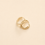 A pair of small gold huggie hoop earrings with a textured, beaded outer edge, displayed on a solid light beige background.
