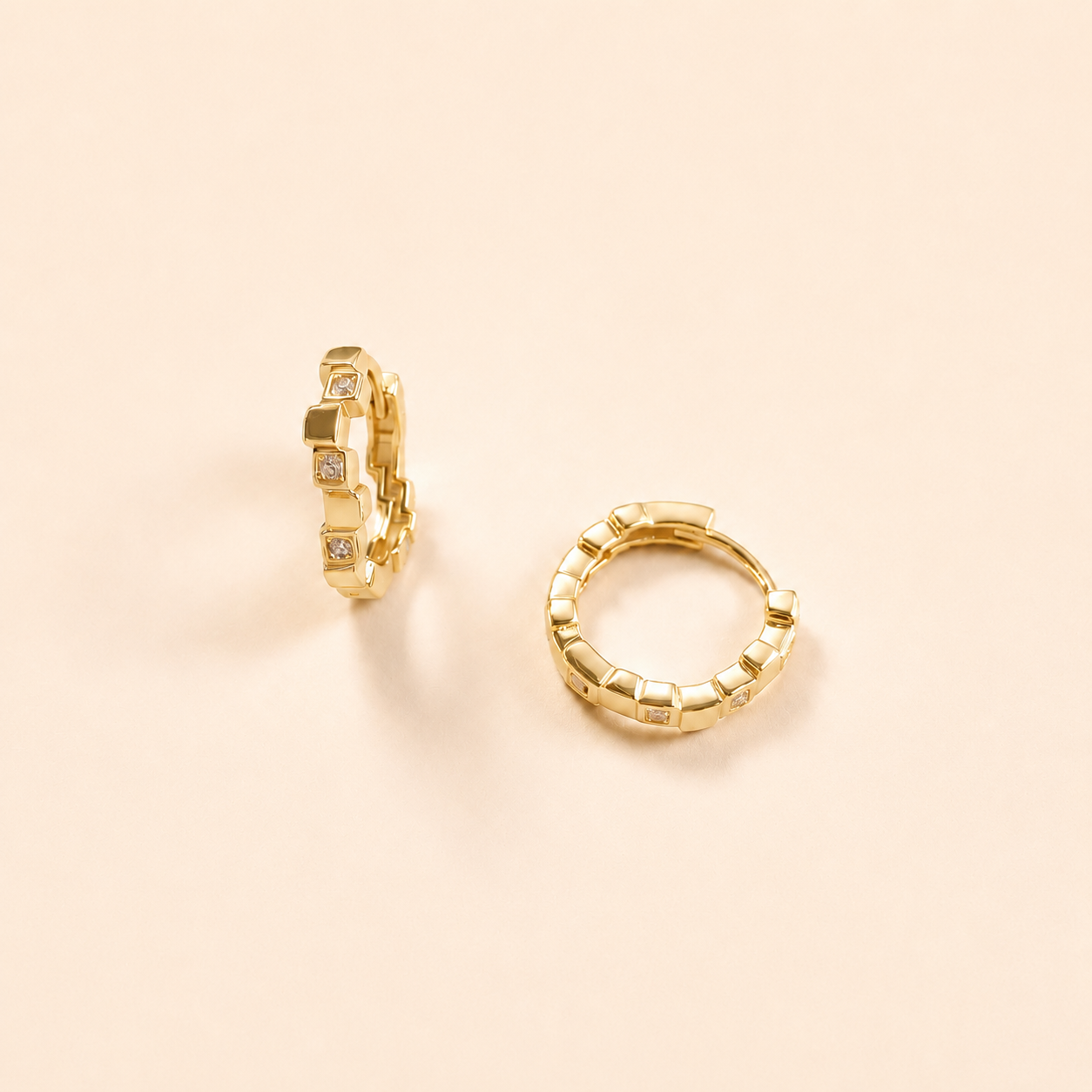 A pair of gold huggie hoop earrings with a textured, block-like segmented design and small clear stones, displayed on a warm beige background.