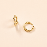 A pair of gold huggie hoop earrings with a textured, block-like segmented design and small clear stones, displayed on a warm beige background.