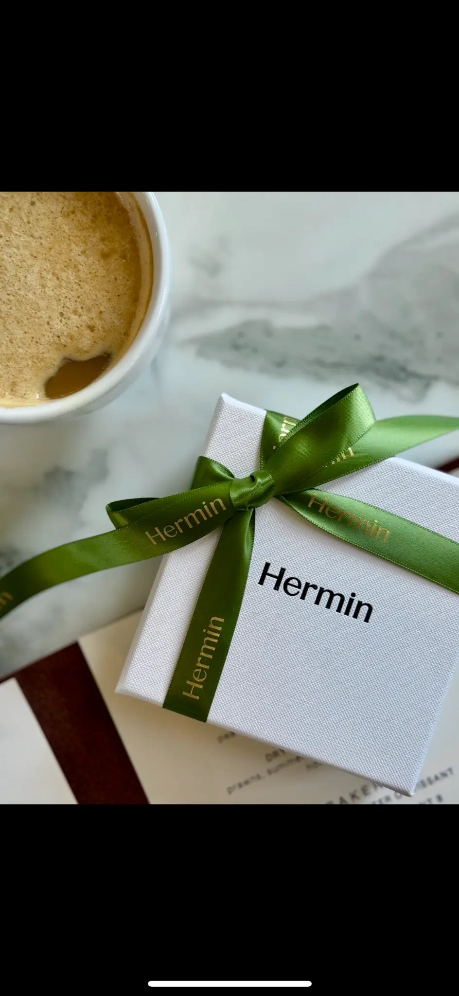 White box with a green ribbon labeled 'Hermin' next to a cup of coffee on a marble surface.